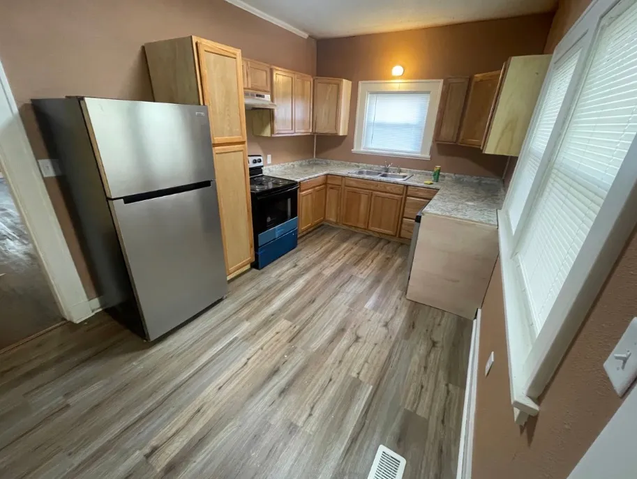 The same kitchen after remodeling, with natural wood cabinets, stainless refrigerator and range, new wood-look plank flooring, and fresh paint