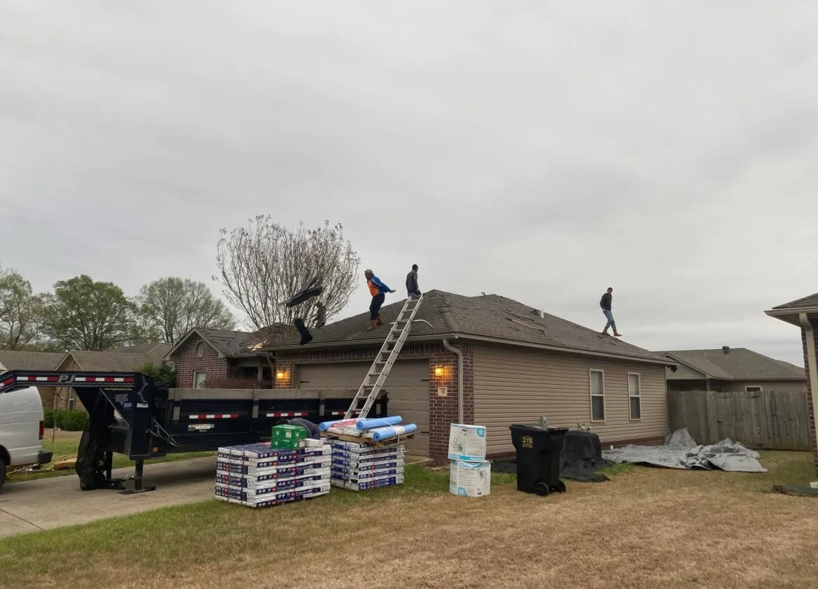 Roofing crew installing a new shingle roof on a home, with shingle bundles staged in the driveway and a work trailer