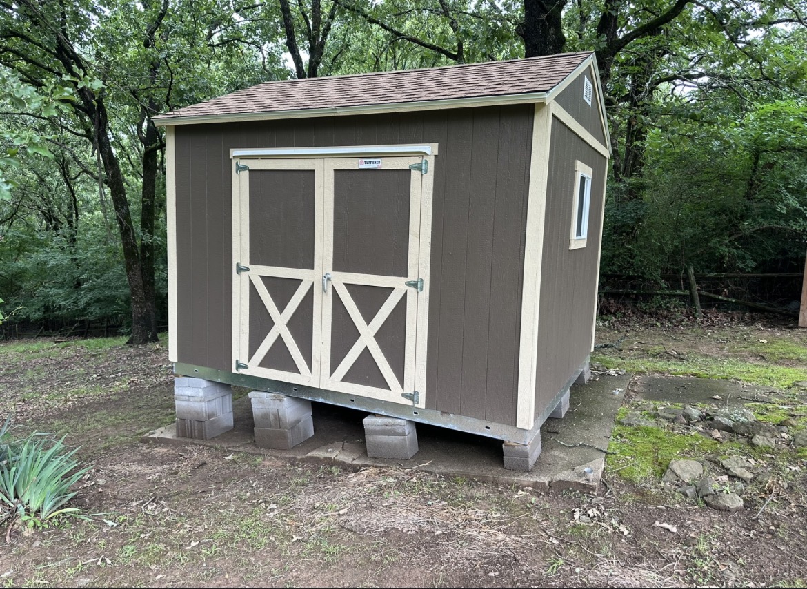 Brown gable-roof shed installed by Renew Home Solutions, with tan trim and double barn-style doors set on a concrete-block foundation among trees