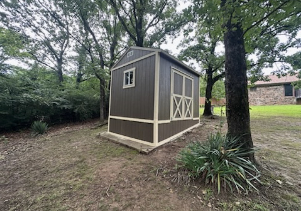 Side view of a brown shed installed by Renew Home Solutions, with tan trim, a window, and double doors in a wooded backyard