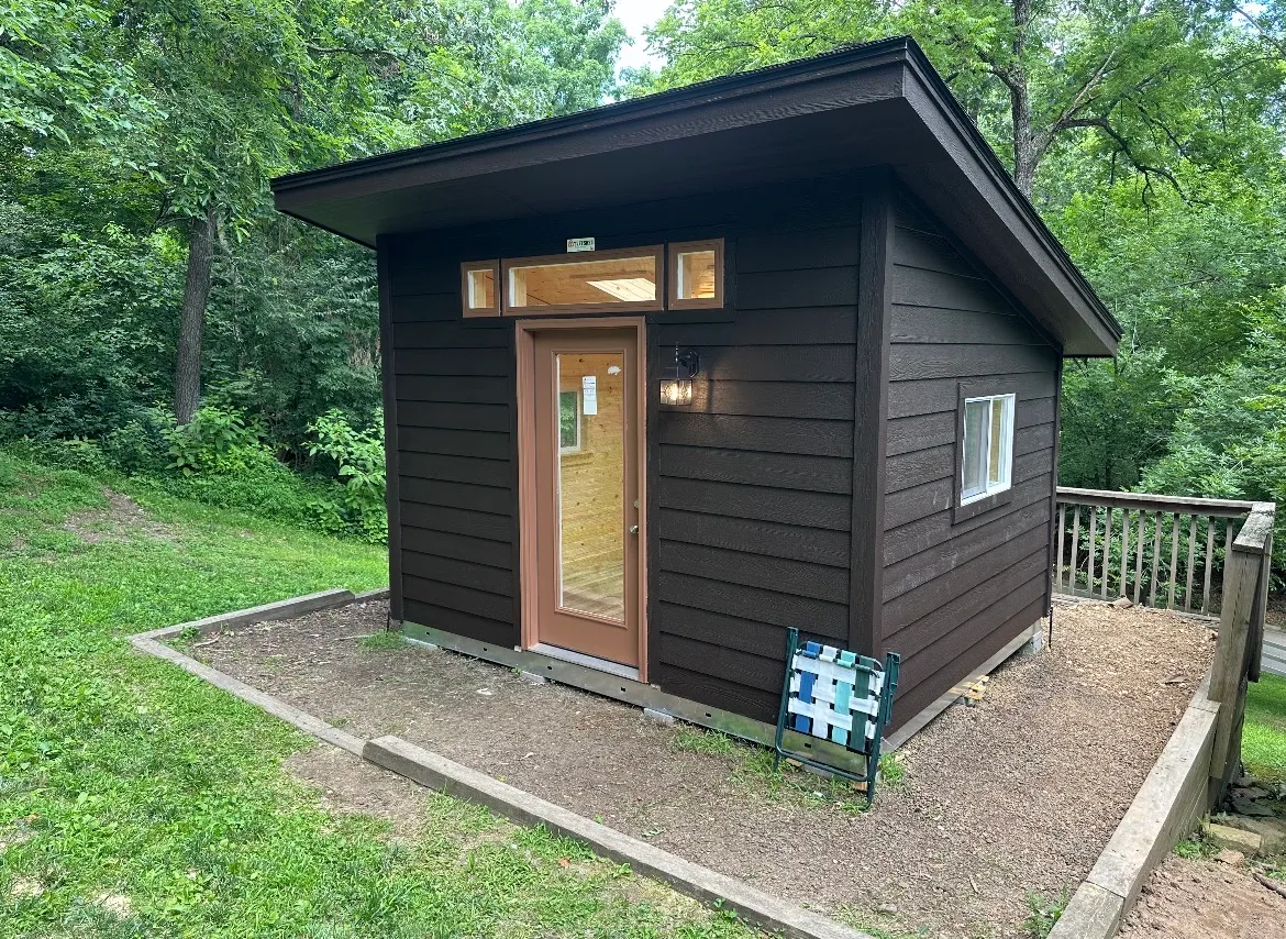 Dark brown modern lean-to shed installed and finished by Renew Home Solutions, with horizontal lap siding, a wood-and-glass entry door, and transom windows on a concrete pad in a wooded Springdale, AR yard