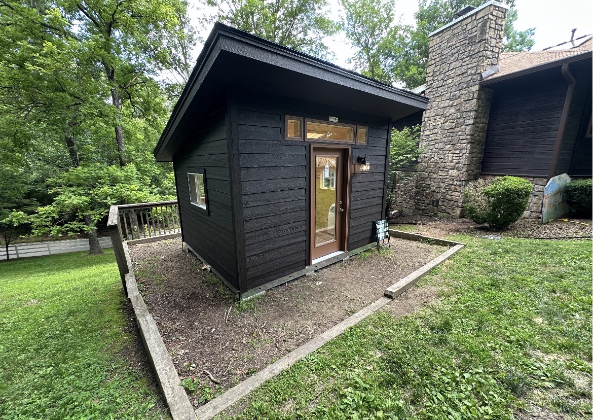Modern lean-to shed finished by Renew Home Solutions with rich black-stained lap siding, a wood-and-glass entry door, and transom windows in a Springdale, AR backyard