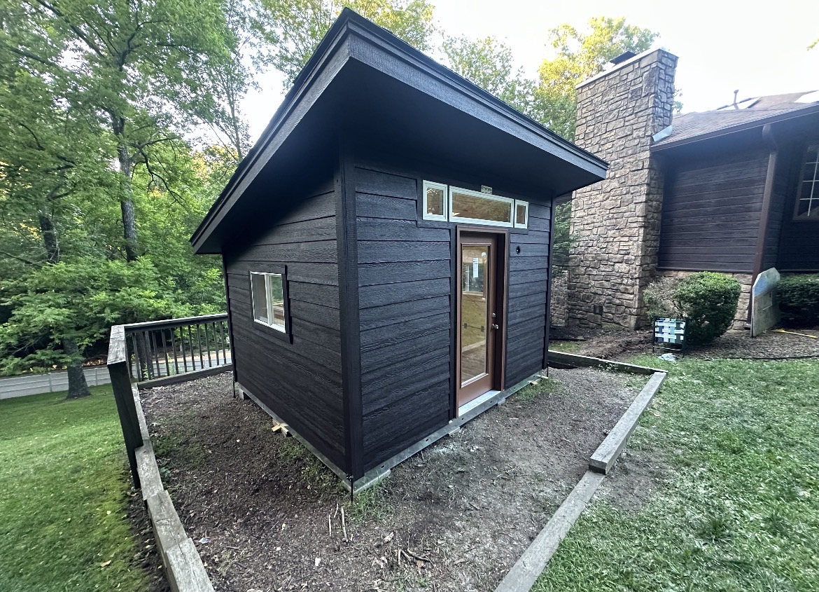 Dark board-and-batten shed installed and finished by Renew Home Solutions, with a glass entry door and transom windows in a Springdale, AR yard