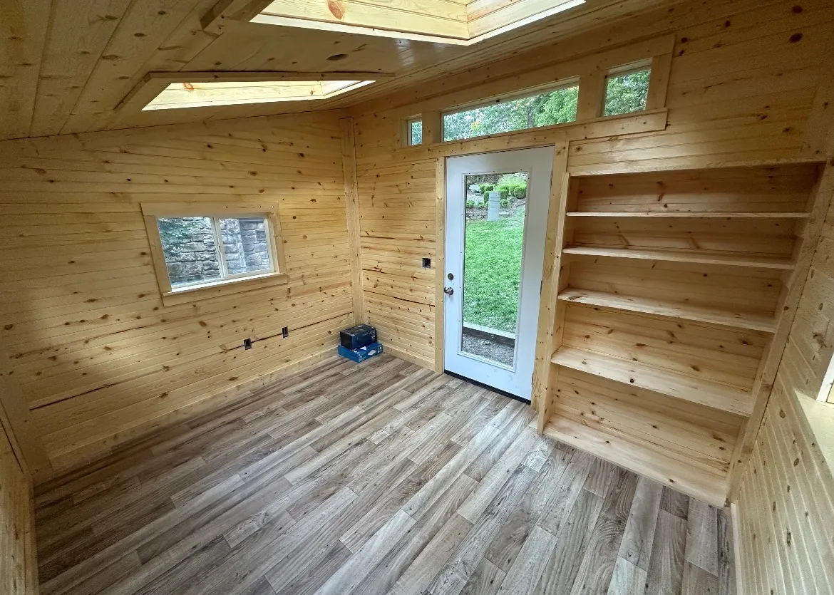 Finished tiny-home-style shed interior with built-in shelving, windows, and wood-plank floors